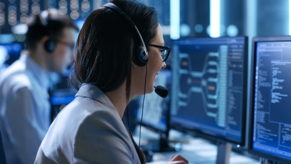 In the System Control Center Woman working in a Technical Support Team Gives Instructions with the Help of the Headsets. Possible Air Traffic/ Power Plant/ Security Room Theme. Mitarbeiterin einer IT-Leasing-Gesellschaft sitzt vor mit dem Rücken zur Kamera gekehrt vor ihrem Bildschirm.